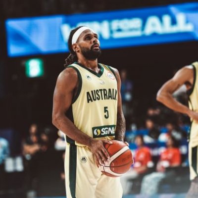 An Indigenous basketball player wearing an Australian uniform holds a basketball and prepares to shoot. There is a teammate and a stadium crowd in the background.